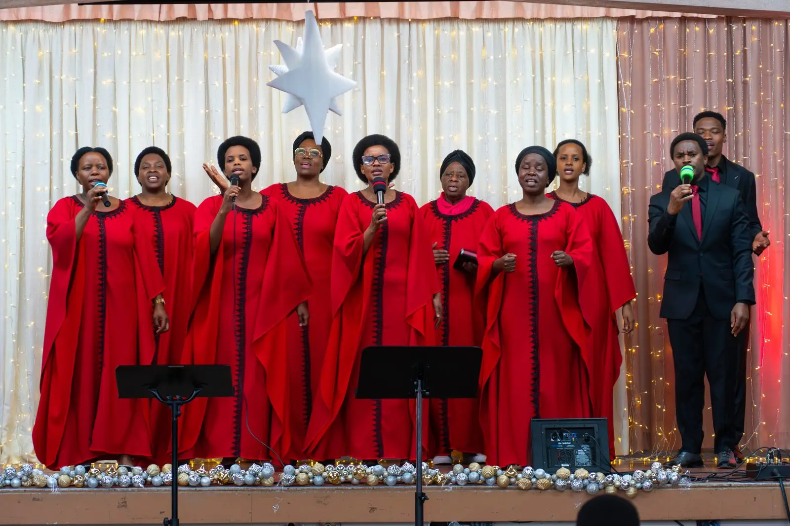A choir of women in flowing red dresses sings on a stage decorated with a white star, led by a man in a black suit.