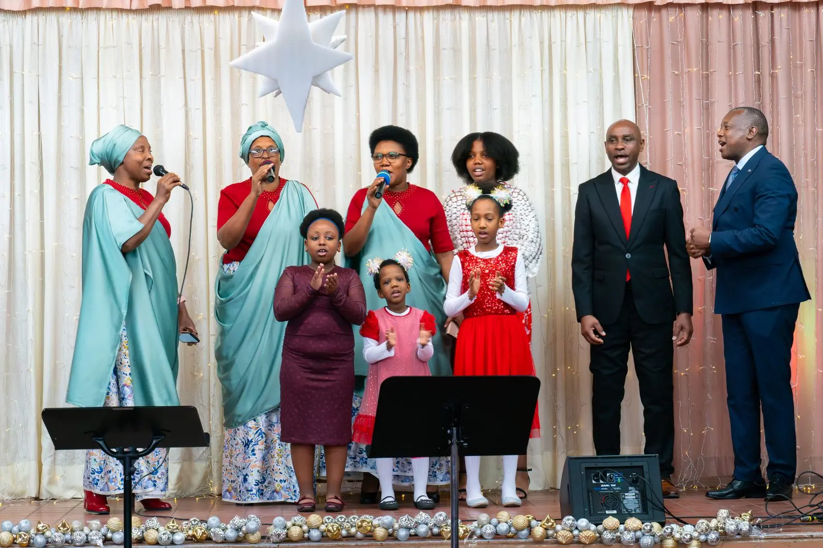 Des femmes en robes turquoise et rouges chantent sur scène avec des fillettes en rouge et deux hommes en complet.