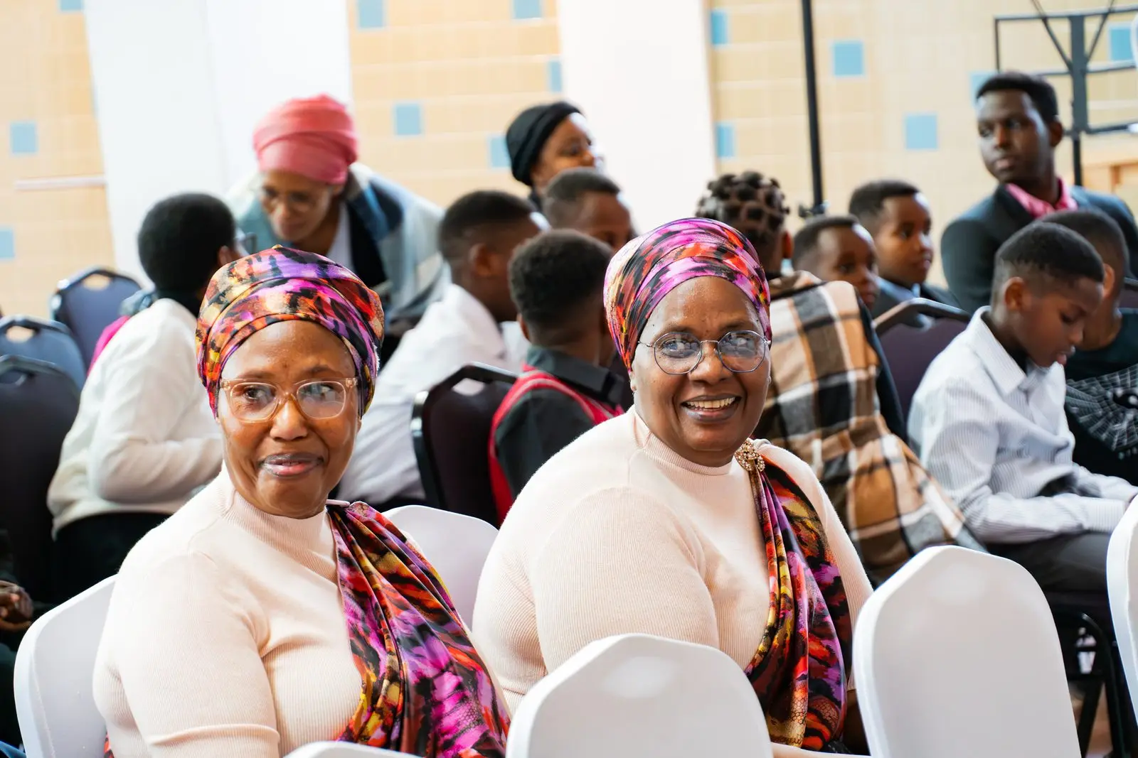 Two smiling women in colourful headwraps seated among the congregation, with children behind them.