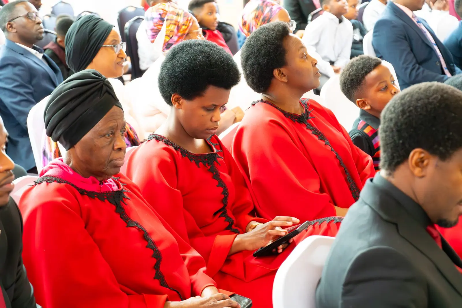 Women in matching red dresses seated in the congregation; one follows along on a tablet.