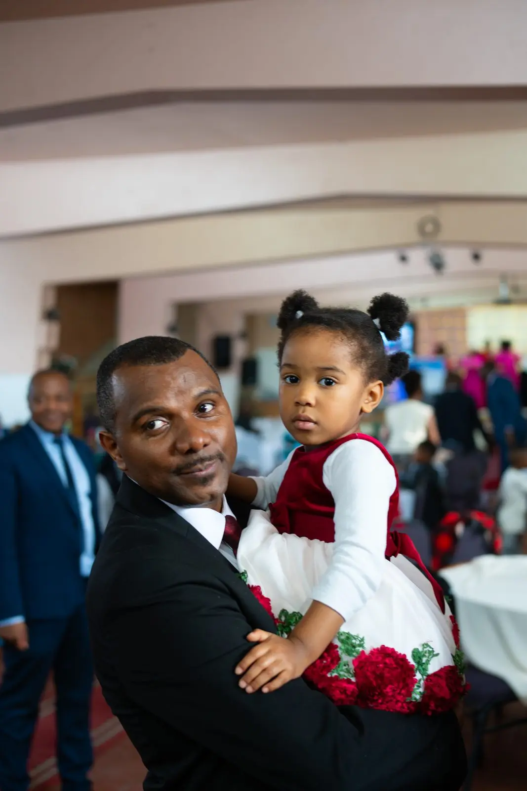Un homme souriant en complet sombre tient une petite fille en robe rouge et blanche, l'assemblée derrière eux.