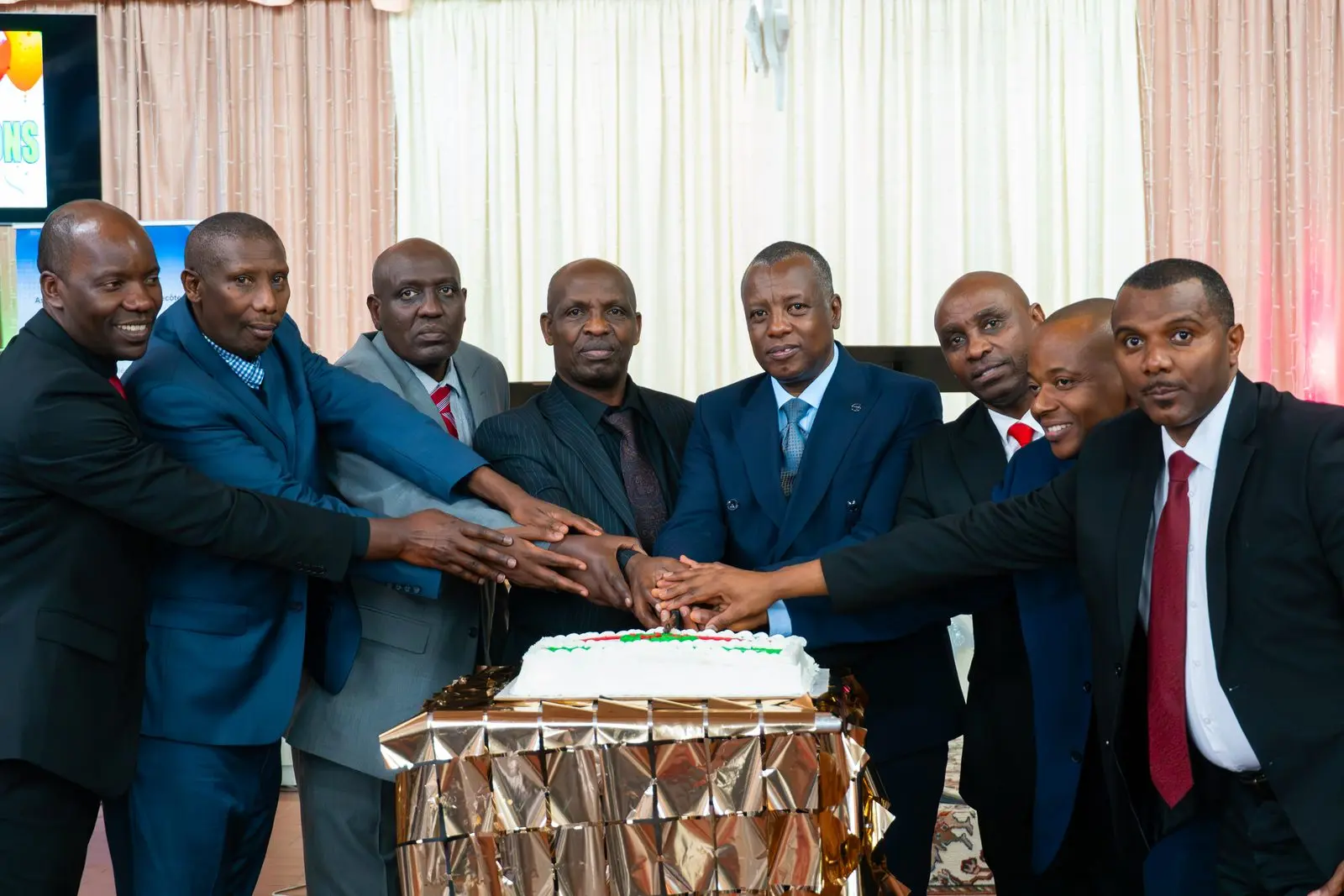 Eight church leaders in suits cut a celebration cake together, every hand on the knife.